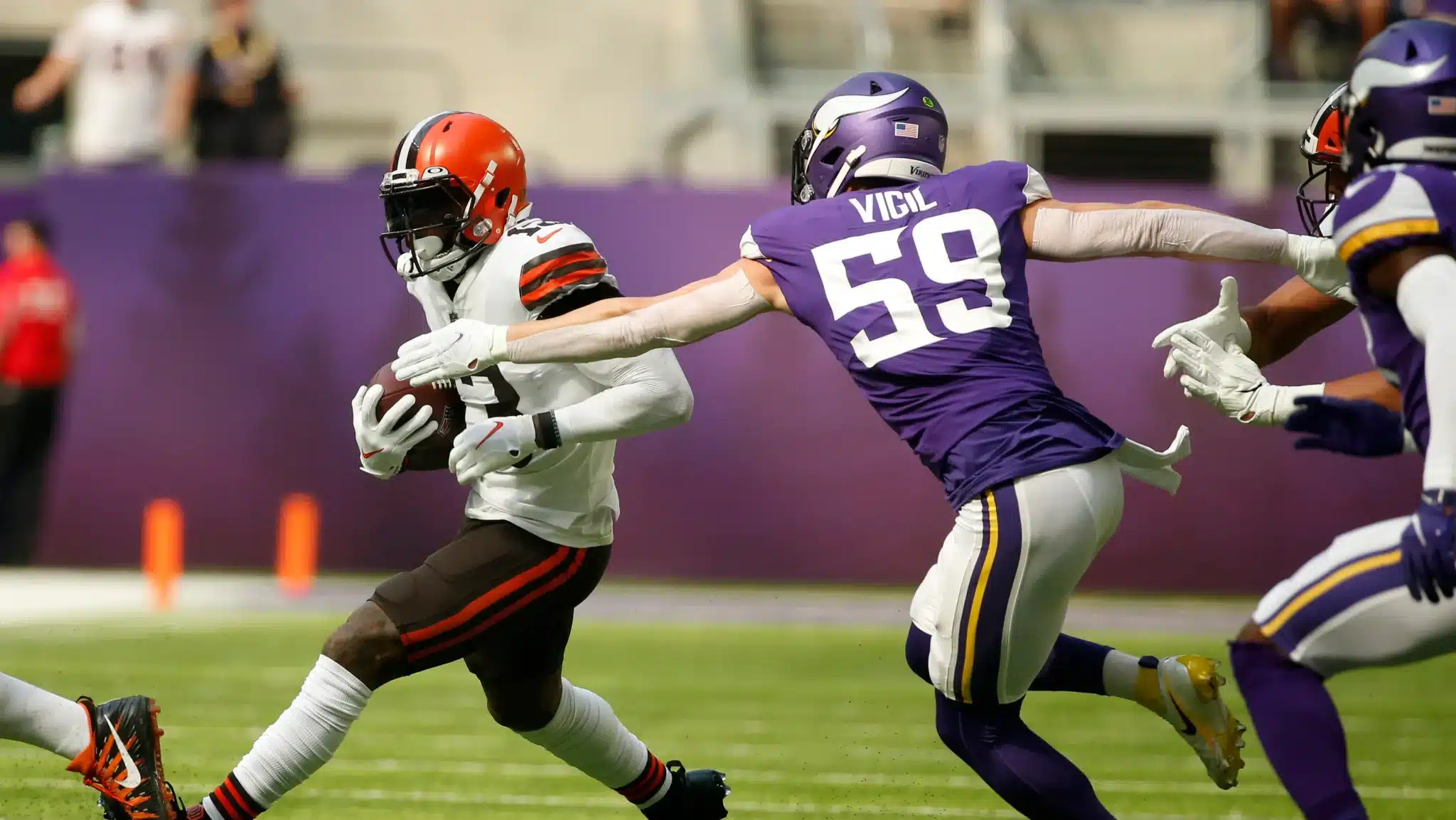 A football player in a white uniform runs with the ball, pursued by a player in a purple uniform with the number 59.