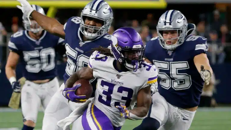 Football player in a purple jersey runs with the ball, pursued by three players in navy blue jerseys on a field.