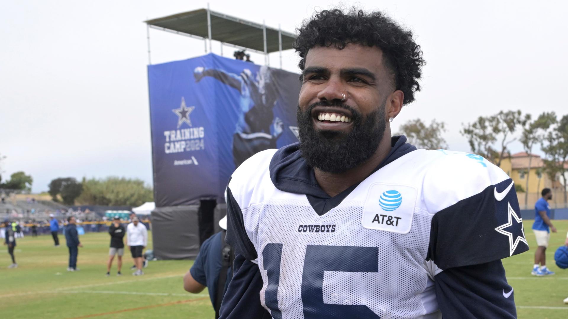 A football player in a Cowboys jersey smiles on a training field with a camp banner in the background.