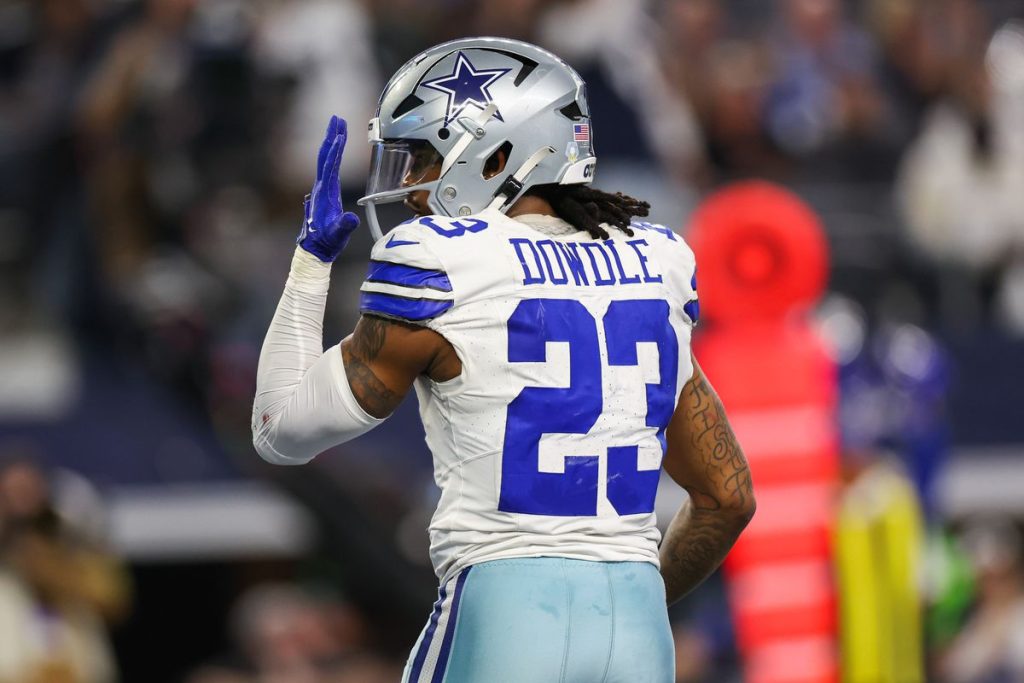 Football player in a white and blue uniform with the name "Dowdle" and number 23 on the jersey, raising his left hand.