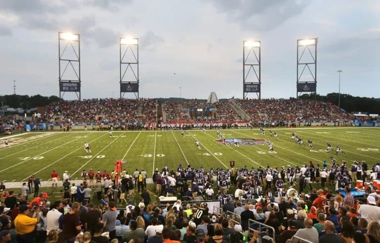 Packed football stadium with fans watching a game under bright lights during twilight, with the field in active play and incorporating recent changes to enhance the experience.