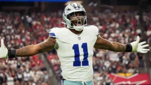 Football player in a white and blue uniform celebrates on the field with arms outstretched during a game.